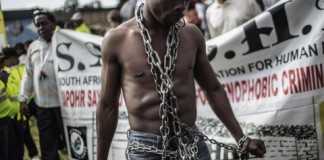 An anti-xenophobia activist stands chained in front of a banner, as thousands of people get ready to march against the recent wave of xenophobic attacks