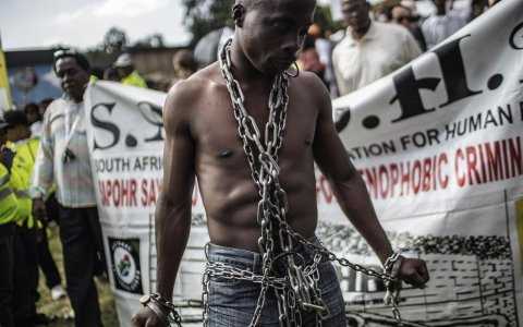 An anti-xenophobia activist stands chained in front of a banner, as thousands of people get ready to march against the recent wave of xenophobic attacks